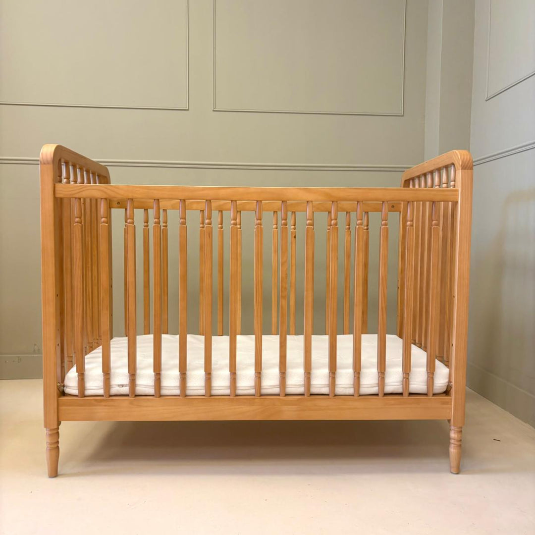 A light natural wood baby cot with vertical slats and a white mattress, photographed in a neutral room with paneled walls.