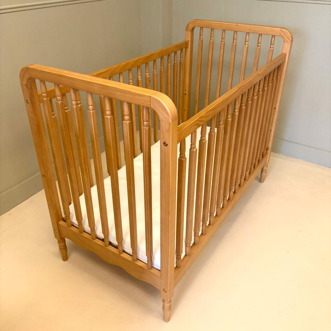 A light natural wood baby crib with turned spindles and a white mattress, photographed in a room with pale paneled walls and a cream floor.