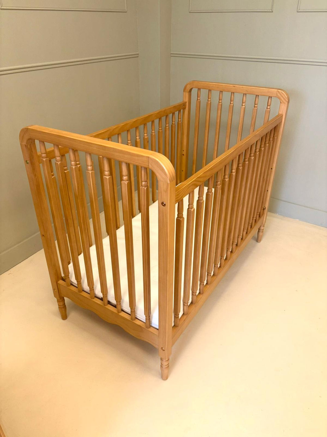 A light natural wood baby cot with turned spindles and a white mattress, photographed in a room with pale paneled walls and a cream floor.